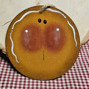 Könnte beinhalten: Ein runder Holzschmuck, der wie ein Lebkuchenmann bemalt ist, mit weißem Zuckerguss und schwarzen Augen. Der Schmuck hat eine Schnur zum Aufhängen.