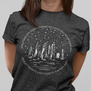 May include: A dark heather grey t-shirt with a white graphic design of a stone circle under a starry sky. The text on the shirt reads "Sometimes you find yourself on a path you never expected. Doesn't mean it can't lead to a bonny place."