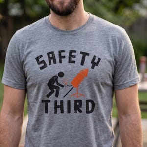K&ouml;nnte beinhalten: Ein graues T-Shirt mit den Worten "SAFETY THIRD" in schwarzen Blockbuchstaben. Das Shirt zeigt eine Grafik einer Person, die eine Rakete anz&uuml;ndet. Die Person tr&auml;gt ein braunes Armband.