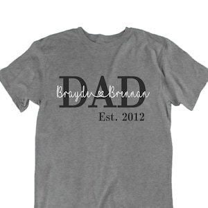 May include: Gray t-shirt with the word "DAD" in large black letters. The text "Brayden & Brennan Est. 2012" is written in white script below the word "DAD".