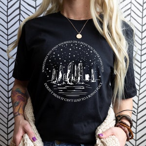 May include: Black t-shirt with a white graphic design featuring a silhouette of a stone circle under a starry sky. The text reads "Sometimes you find yourself on a path you never expected. Doesn't mean it can't lead to a bonny place."