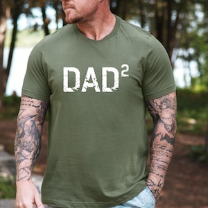 May include: Olive green t-shirt with the white text "DAD²" in a distressed font. The shirt has a crew neck and short sleeves. The image is taken outdoors with a blurred background of trees.