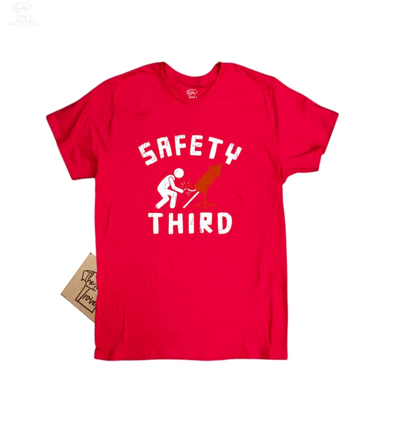 K&ouml;nnte beinhalten: Ein leuchtend rotes T-Shirt mit dem wei&szlig;en Schriftzug "SAFETY THIRD", zusammen mit einer Strichm&auml;nnchen-Figur und einer Rakete. Ein kleines Etikett ist angebracht. Es ist ein Freizeitkleidungsst&uuml;ck.
