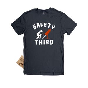 K&ouml;nnte beinhalten: Dunkelgraues T-Shirt mit den Worten "SAFETY THIRD" in wei&szlig;en Blockbuchstaben. Das Shirt zeigt eine Strichm&auml;nnchen, das ein rotes Feuerwerk anz&uuml;ndet. Ein kleines braunes Etikett ist am Shirt befestigt.