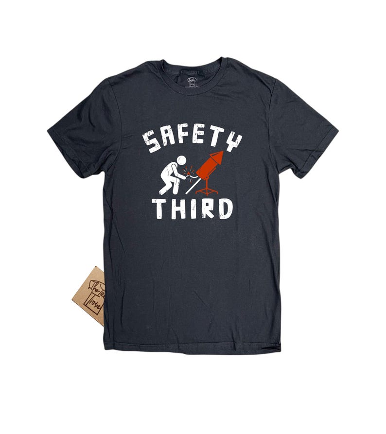 K&ouml;nnte beinhalten: Dunkelgraues T-Shirt mit den Worten "SAFETY THIRD" in wei&szlig;en Blockbuchstaben. Das Shirt zeigt eine Strichm&auml;nnchen, das ein rotes Feuerwerk anz&uuml;ndet. Ein kleines braunes Etikett ist am Shirt befestigt.