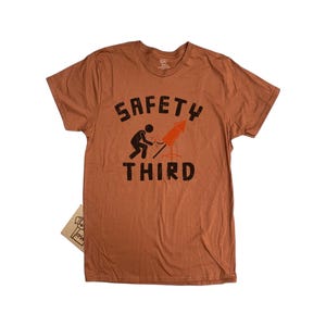 K&ouml;nnte beinhalten: Ein rostfarbenes T-Shirt mit dem schwarzen Schriftzug "SAFETY THIRD". Eine Grafik zeigt eine Person, die eine Rakete anz&uuml;ndet. Ein kleines Etikett ist am T-Shirt befestigt.