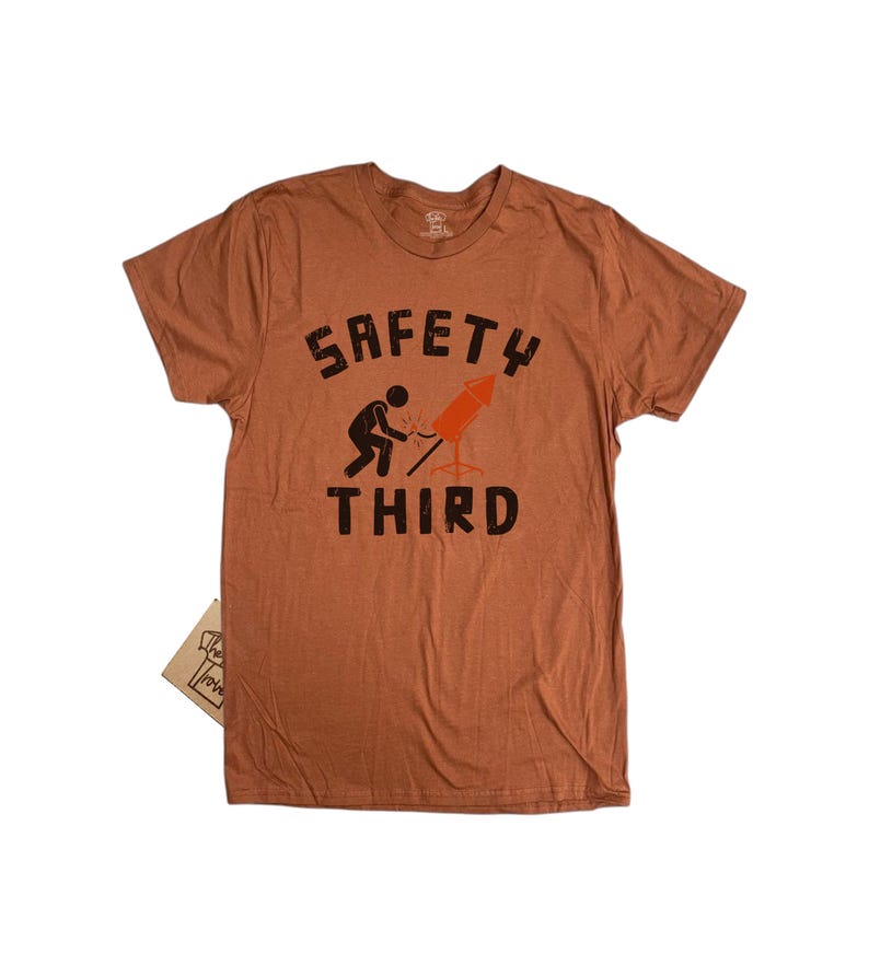 K&ouml;nnte beinhalten: Ein rostfarbenes T-Shirt mit dem schwarzen Schriftzug "SAFETY THIRD". Eine Grafik zeigt eine Person, die eine Rakete anz&uuml;ndet. Ein kleines Etikett ist am T-Shirt befestigt.