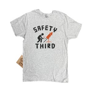 K&ouml;nnte beinhalten: Hellgraues T-Shirt mit dem Text "SAFETY THIRD" in schwarzer, verwaschener Schrift. Eine Grafik zeigt eine Strichm&auml;nnchen, das eine Rakete schwei&szlig;t. Die Rakete ist orange und rot. Ein kleines braunes Etikett ist angebracht.