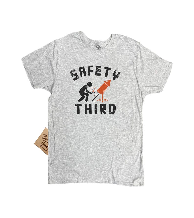 K&ouml;nnte beinhalten: Hellgraues T-Shirt mit dem Text "SAFETY THIRD" in schwarzer, verwaschener Schrift. Eine Grafik zeigt eine Strichm&auml;nnchen, das eine Rakete schwei&szlig;t. Die Rakete ist orange und rot. Ein kleines braunes Etikett ist angebracht.