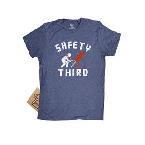 K&ouml;nnte beinhalten: Ein blau meliertes T-Shirt mit dem wei&szlig;en Schriftzug "SAFETY THIRD". Das Shirt zeigt eine Grafik einer Strichm&auml;nnchen, das ein rotes Feuerwerk anz&uuml;ndet. Ein kleines braunes Pappschild ist sichtbar.