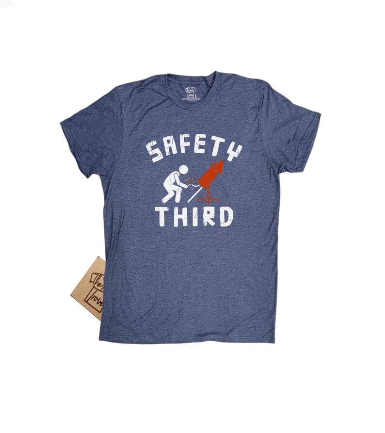 K&ouml;nnte beinhalten: Ein blau meliertes T-Shirt mit dem wei&szlig;en Schriftzug "SAFETY THIRD". Das Shirt zeigt eine Grafik einer Strichm&auml;nnchen, das ein rotes Feuerwerk anz&uuml;ndet. Ein kleines braunes Pappschild ist sichtbar.