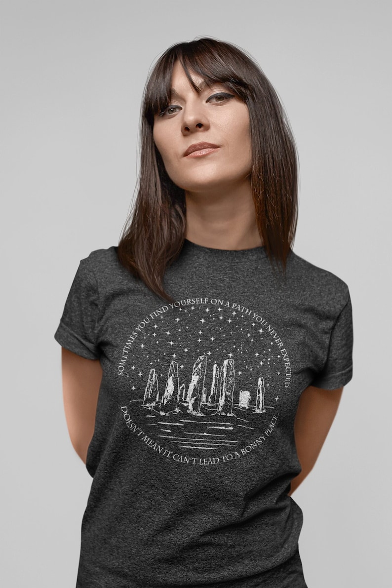May include: A dark heather grey t-shirt with a white graphic design of a stone circle under a starry sky. The text on the shirt reads "Sometimes you find yourself on a path you never expected. Doesn't mean it can't lead to a bonny place."