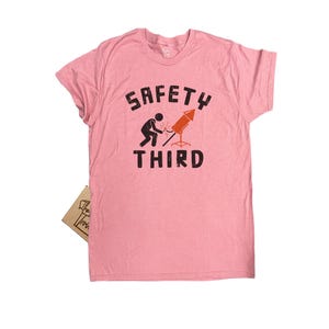 K&ouml;nnte beinhalten: Hellrosa T-Shirt mit dem Text "SAFETY THIRD" in schwarzen Buchstaben. Unter dem Wort "SAFETY" ist eine Grafik einer Person abgebildet, die eine Rakete anz&uuml;ndet. Das T-Shirt hat kurze &Auml;rmel und einen Rundhalsausschnitt.