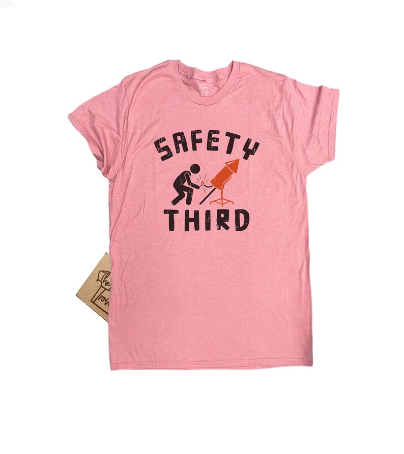 K&ouml;nnte beinhalten: Hellrosa T-Shirt mit dem Text "SAFETY THIRD" in schwarzen Buchstaben. Unter dem Wort "SAFETY" ist eine Grafik einer Person abgebildet, die eine Rakete anz&uuml;ndet. Das T-Shirt hat kurze &Auml;rmel und einen Rundhalsausschnitt.
