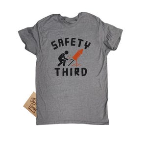K&ouml;nnte beinhalten: Graues T-Shirt mit dem Text "SAFETY THIRD" in verblichener Schrift. Die Grafik zeigt eine Strichm&auml;nnchen, das eine Rakete schwei&szlig;t. Ein kleines braunes Etikett ist am T-Shirt befestigt.