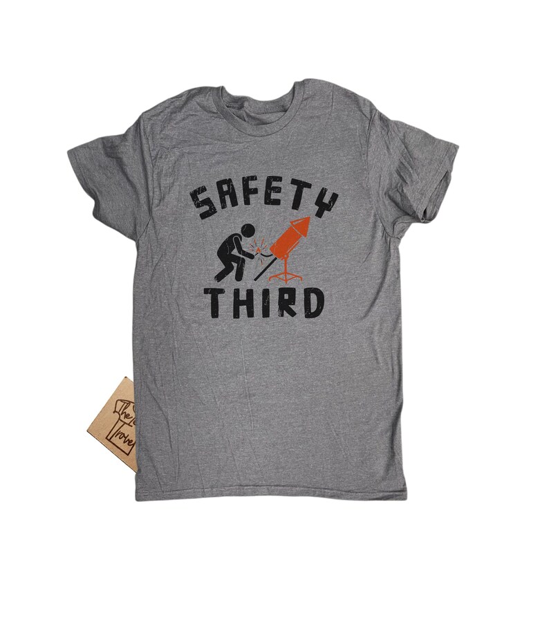 K&ouml;nnte beinhalten: Graues T-Shirt mit dem Text "SAFETY THIRD" in verblichener Schrift. Die Grafik zeigt eine Strichm&auml;nnchen, das eine Rakete schwei&szlig;t. Ein kleines braunes Etikett ist am T-Shirt befestigt.