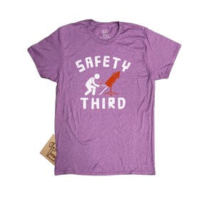 K&ouml;nnte beinhalten: Ein meliertes lila T-Shirt mit dem wei&szlig;en Schriftzug &bdquo;SAFETY THIRD&ldquo;. Das T-Shirt zeigt eine Grafik einer Person, die ein rotes Feuerwerk anz&uuml;ndet. Ein kleines Etikett ist am T-Shirt befestigt.