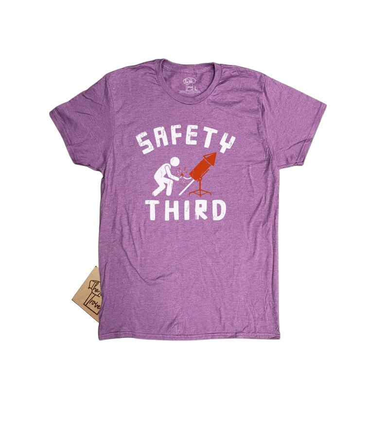 K&ouml;nnte beinhalten: Ein meliertes lila T-Shirt mit dem wei&szlig;en Schriftzug &bdquo;SAFETY THIRD&ldquo;. Das T-Shirt zeigt eine Grafik einer Person, die ein rotes Feuerwerk anz&uuml;ndet. Ein kleines Etikett ist am T-Shirt befestigt.