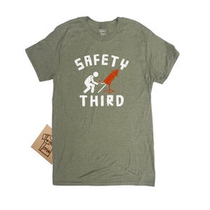 K&ouml;nnte beinhalten: Olivgr&uuml;nes T-Shirt mit dem wei&szlig;en Text "SAFETY THIRD". Die Grafik zeigt eine Strichm&auml;nnchen, das eine rote Rakete anz&uuml;ndet. Ein kleines braunes Etikett ist am T-Shirt befestigt.