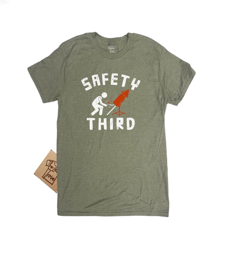 K&ouml;nnte beinhalten: Olivgr&uuml;nes T-Shirt mit dem wei&szlig;en Text "SAFETY THIRD". Die Grafik zeigt eine Strichm&auml;nnchen, das eine rote Rakete anz&uuml;ndet. Ein kleines braunes Etikett ist am T-Shirt befestigt.