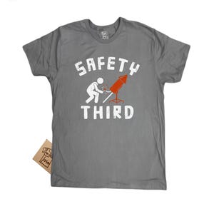 K&ouml;nnte beinhalten: Graues T-Shirt mit dem wei&szlig;en Schriftzug "SAFETY THIRD". Das Design zeigt eine Strichm&auml;nnchenfigur, die ein rotes Feuerwerk anz&uuml;ndet. Ein kleines braunes Etikett ist am T-Shirt befestigt.