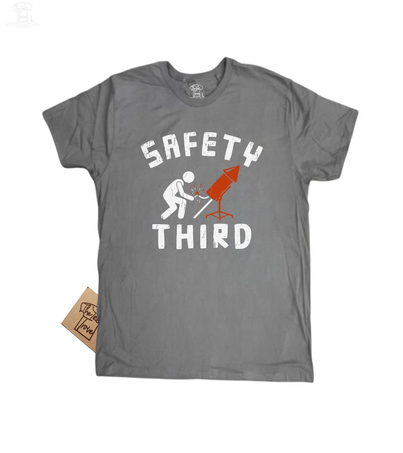 K&ouml;nnte beinhalten: Graues T-Shirt mit dem wei&szlig;en Schriftzug "SAFETY THIRD". Das Design zeigt eine Strichm&auml;nnchenfigur, die ein rotes Feuerwerk anz&uuml;ndet. Ein kleines braunes Etikett ist am T-Shirt befestigt.