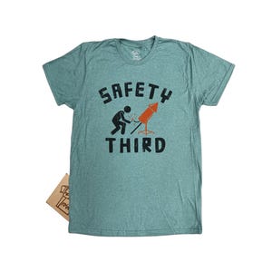 K&ouml;nnte beinhalten: Ein t&uuml;rkisfarbenes T-Shirt mit den Worten "SAFETY THIRD" in schwarzen Blockbuchstaben. Eine Strichm&auml;nnchen z&uuml;ndet eine rote und orangefarbene Feuerwerksrakete an. Das Shirt ist aus weichem Material.