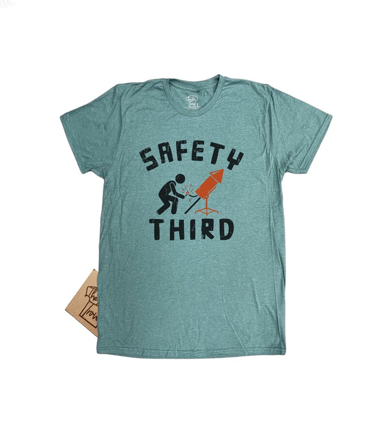 K&ouml;nnte beinhalten: Ein t&uuml;rkisfarbenes T-Shirt mit den Worten "SAFETY THIRD" in schwarzen Blockbuchstaben. Eine Strichm&auml;nnchen z&uuml;ndet eine rote und orangefarbene Feuerwerksrakete an. Das Shirt ist aus weichem Material.