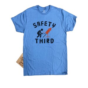 K&ouml;nnte beinhalten: Hellblaues T-Shirt mit dem Text "SAFETY THIRD" in schwarzen Buchstaben. Die Grafik zeigt eine Strichm&auml;nnchen, das ein rotes Feuerwerk anz&uuml;ndet. Ein kleines braunes Pappschild ist unten sichtbar.