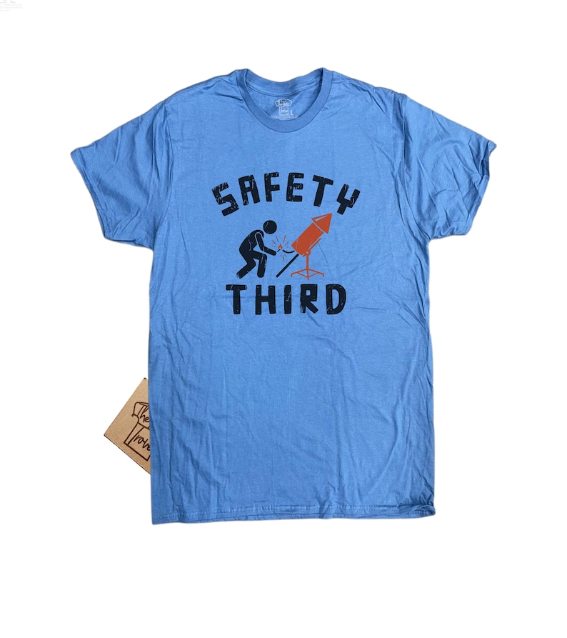 K&ouml;nnte beinhalten: Hellblaues T-Shirt mit dem Text "SAFETY THIRD" in schwarzen Buchstaben. Die Grafik zeigt eine Strichm&auml;nnchen, das ein rotes Feuerwerk anz&uuml;ndet. Ein kleines braunes Pappschild ist unten sichtbar.