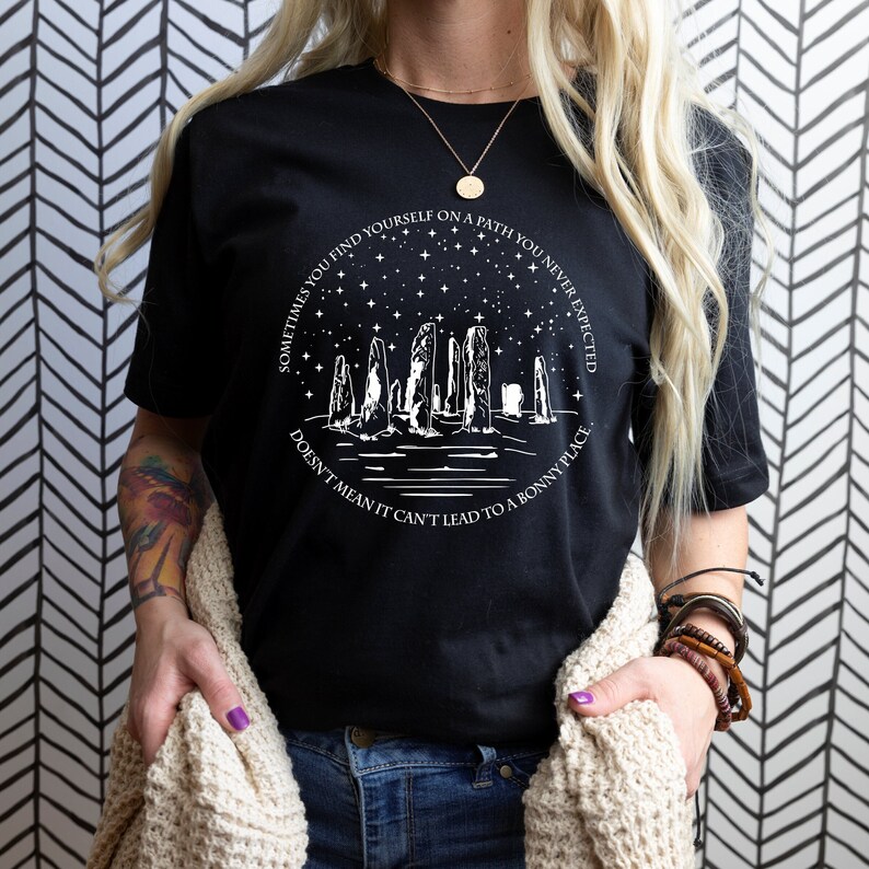 May include: Black t-shirt with a white graphic design featuring a silhouette of a stone circle under a starry sky. The text reads "Sometimes you find yourself on a path you never expected. Doesn't mean it can't lead to a bonny place."