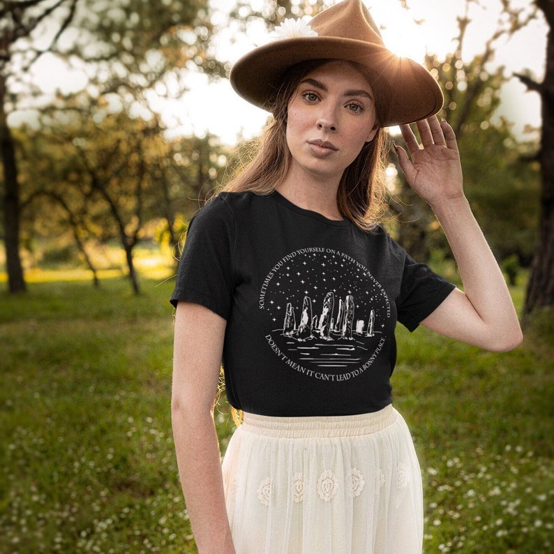 May include: A black t-shirt with a white graphic design of a circle with stars and a line of standing stones. The text on the shirt reads 'Sometimes you find yourself on a path you never expected. Doesn't mean it can't lead to a pretty place.'