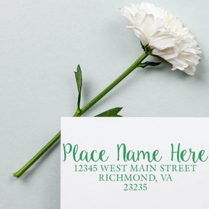 May include: A white card with green text that reads "Place Name Here", "12345 West Main Street", "Richmond, VA", and "23235". A white flower with green stem is laying on the card.