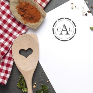 May include: A white piece of paper with a black and white circle stamp that says "CAL" in a decorative font. The paper is on a black surface with a wooden spoon, red and white checkered fabric, and spices.