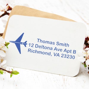 Address stamp featuring an airplane design, perfect for air travel enthusiasts. The address label displays Thomas Smith, 12 Deltona Ave Apt B, Richmond, VA 23230 on a card, surrounded by delicate white flowers.