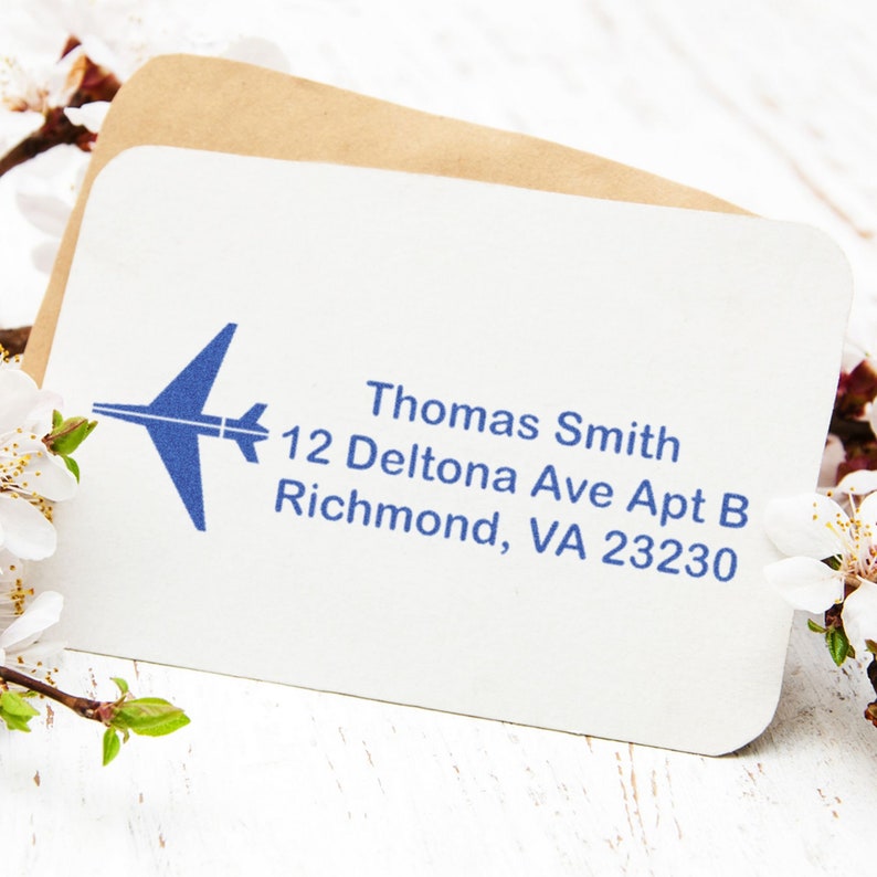 Address stamp featuring an airplane design, perfect for air travel enthusiasts. The address label displays Thomas Smith, 12 Deltona Ave Apt B, Richmond, VA 23230 on a card, surrounded by delicate white flowers.