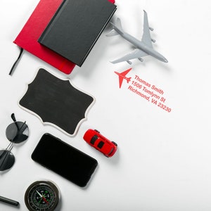 A personalized gift featuring an ink stamp with an airplane design, perfect for an airplane address stamp. Includes a red notebook, toy car, and compass on a white background. Ideal for travel enthusiasts.