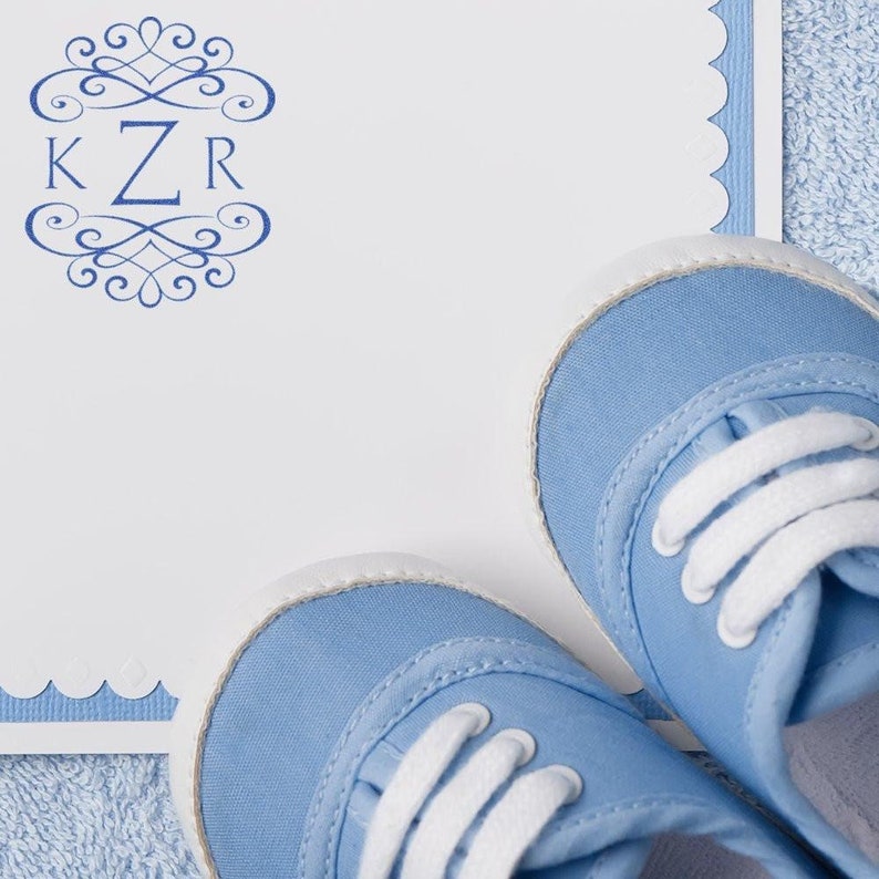 Blue baby shoes next to a personalized stamp on a white card, perfect for wedding DIY projects. The ink stamp features elegant initials, adding a custom touch to invitations or thank you notes. Ideal for creative, personalized wedding crafts.