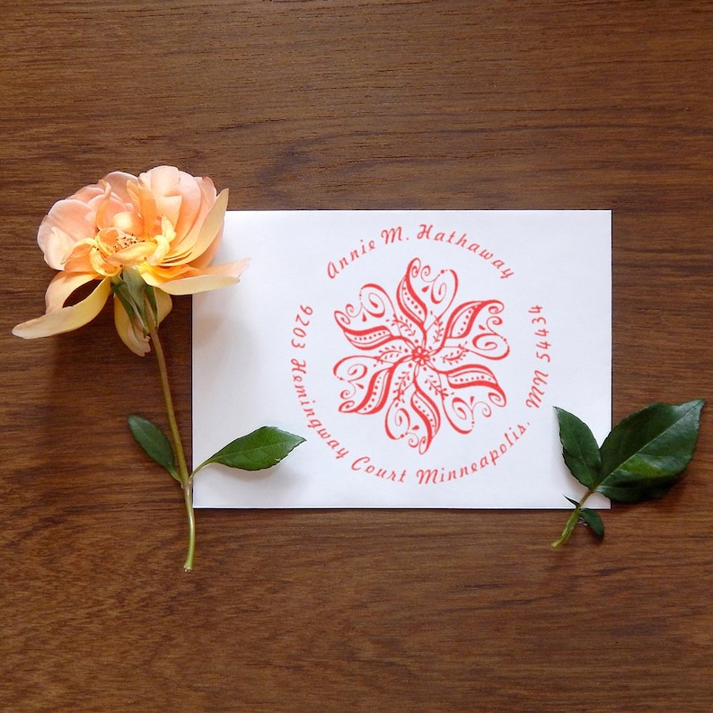 A self-inking address stamp featuring a floral design is displayed on an envelope, accompanied by a flower rubber stamp. The intricate pattern adds elegance to personalized correspondence.