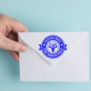 Hand holding an envelope with a blue Christmas Tag Stamp reading Sleigh Mail The North Pole on a light blue background. Ideal for craft supplies, stamping projects, and stamping ideas for festive occasions.
