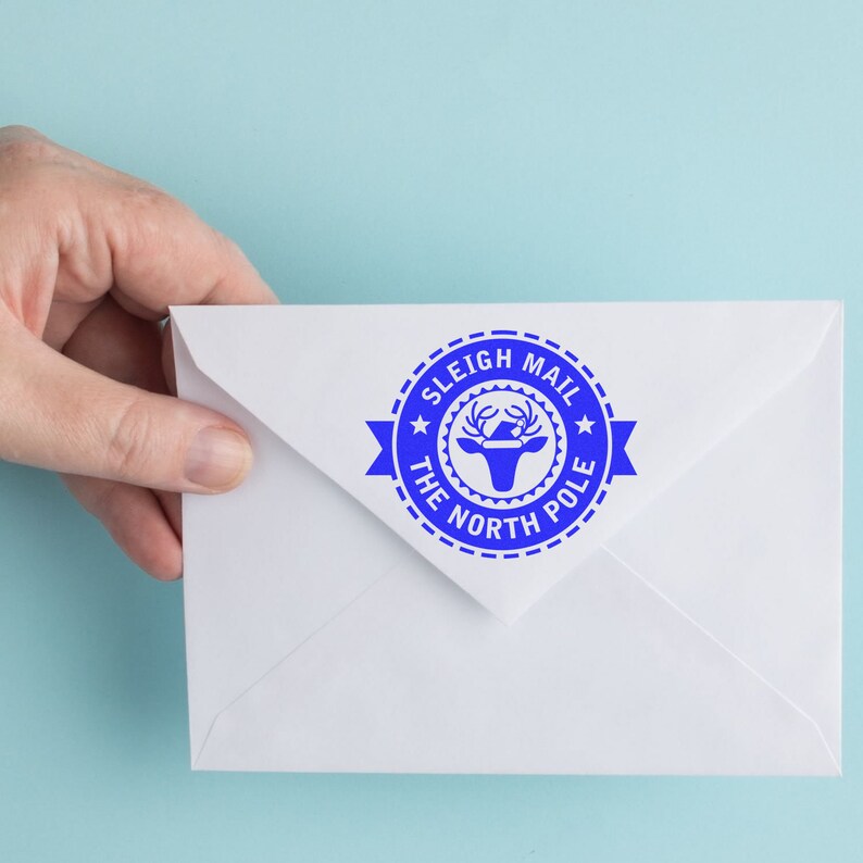 Hand holding an envelope with a blue Christmas Tag Stamp reading Sleigh Mail The North Pole on a light blue background. Ideal for craft supplies, stamping projects, and stamping ideas for festive occasions.