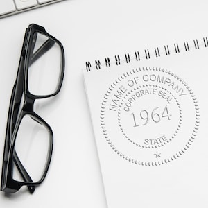 May include: Black-framed eyeglasses and a spiral-bound notepad with a corporate seal design. The seal includes the text "NAME OF COMPANY", "CORPORATE SEAL", "1964", and "STATE". The items are on a white surface.