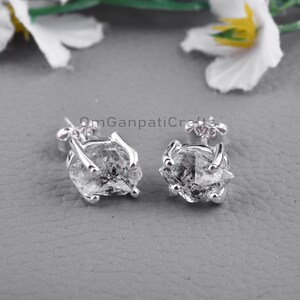 May include: A pair of silver stud earrings featuring rough-cut, clear quartz crystals. The earrings have a claw-like setting, and the posts are adorned with a floral design. The background is a neutral gray.