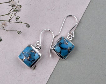 May include: A pair of square drop earrings with a silver frame. The earrings feature a turquoise stone with gold flecks. The earrings are displayed on a light background with a sprig of white flowers.
