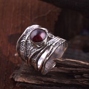 May include: A silver ring featuring an elaborate design and a prominent, circular, dark red gemstone. The ring showcases intricate patterns and textures, with a polished surface. The gemstone is centrally set, drawing attention to the ring's focal point.