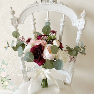 May include: A bridal bouquet featuring burgundy, blush pink, and white roses, eucalyptus foliage, and burgundy berries. The bouquet is tied with a white satin ribbon.