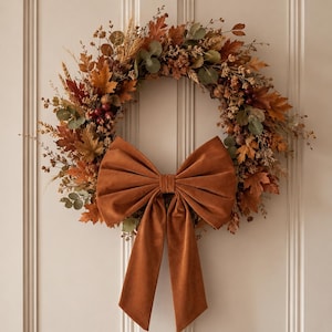 May include: A circular wreath featuring a mix of dried autumn leaves in shades of orange, brown, and muted green, accented with small berries and wheat stalks. A large, solid burnt orange velvet bow is attached to the bottom centre of the wreath.