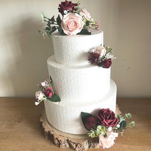 May include: A three-tiered white cake adorned with artificial flowers in pink, burgundy, and green. The cake rests on a rustic wooden base, creating a charming and elegant display. Ideal for weddings or special occasions.