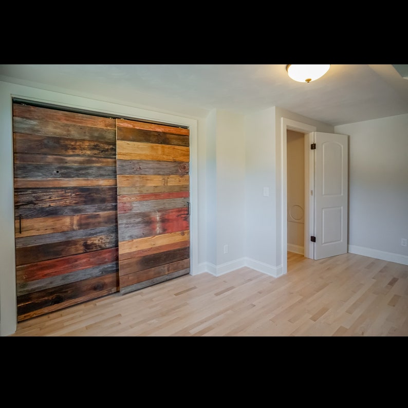 Puede incluir: Un dormitorio con suelo de madera clara y paredes blancas. El punto focal es un juego de puertas correderas de armario hechas de tablones de madera recuperada en varios colores, incluyendo marr&oacute;n, rojo y naranja. Una puerta blanca est&aacute; entreabierta en el fondo.