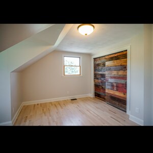 Puede incluir: Vista interior de una habitaci&oacute;n con suelo de madera clara y techo inclinado. Una peque&ntilde;a ventana est&aacute; centrada en la pared, y una gran puerta corredera de granero de madera reciclada est&aacute; a la derecha. Una l&aacute;mpara est&aacute; montada en el techo.